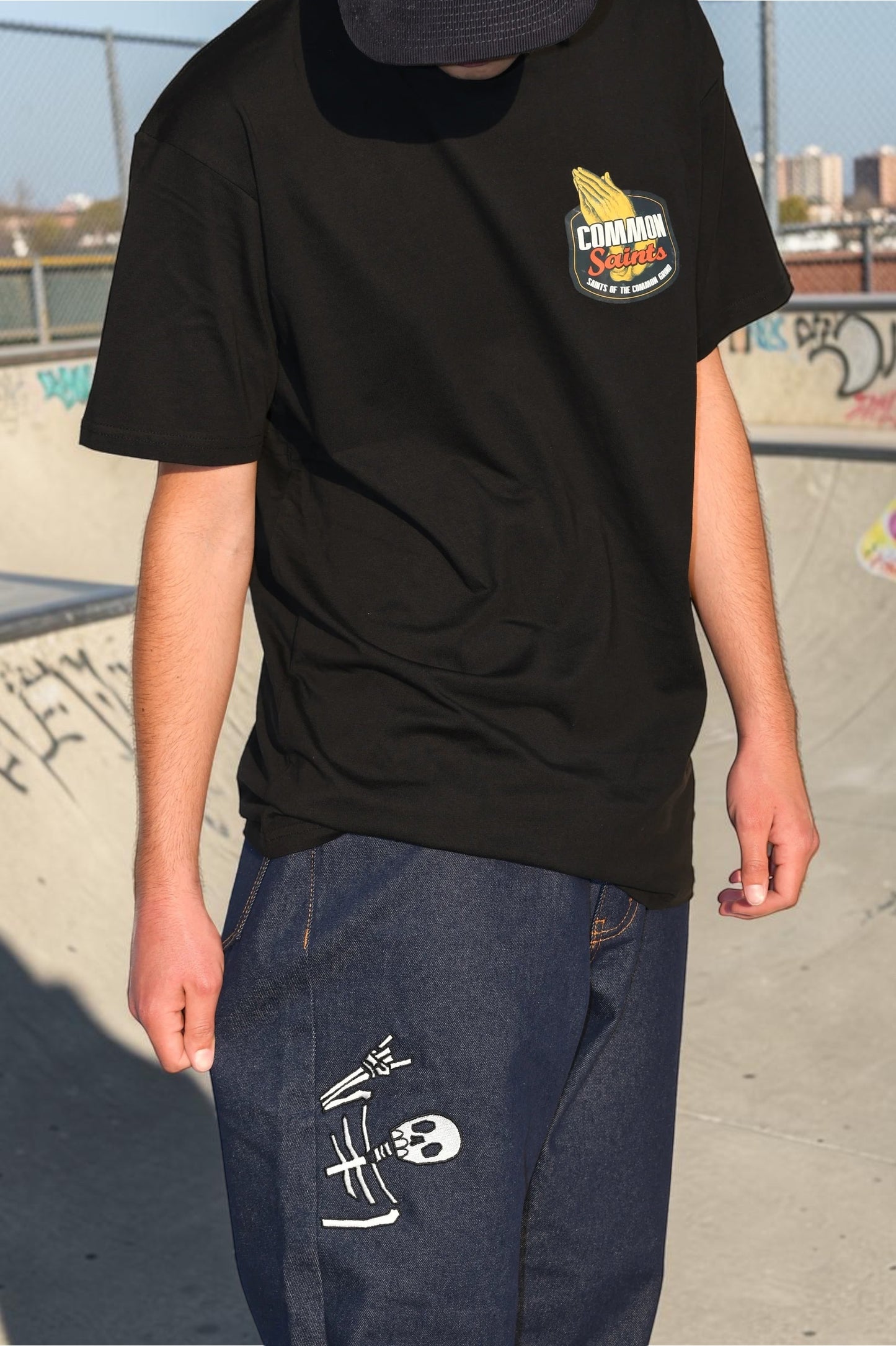 Model wearing Common Saints dark indigo jeans with white embroidered skull graphic on the left thigh at an outdoor skatepark