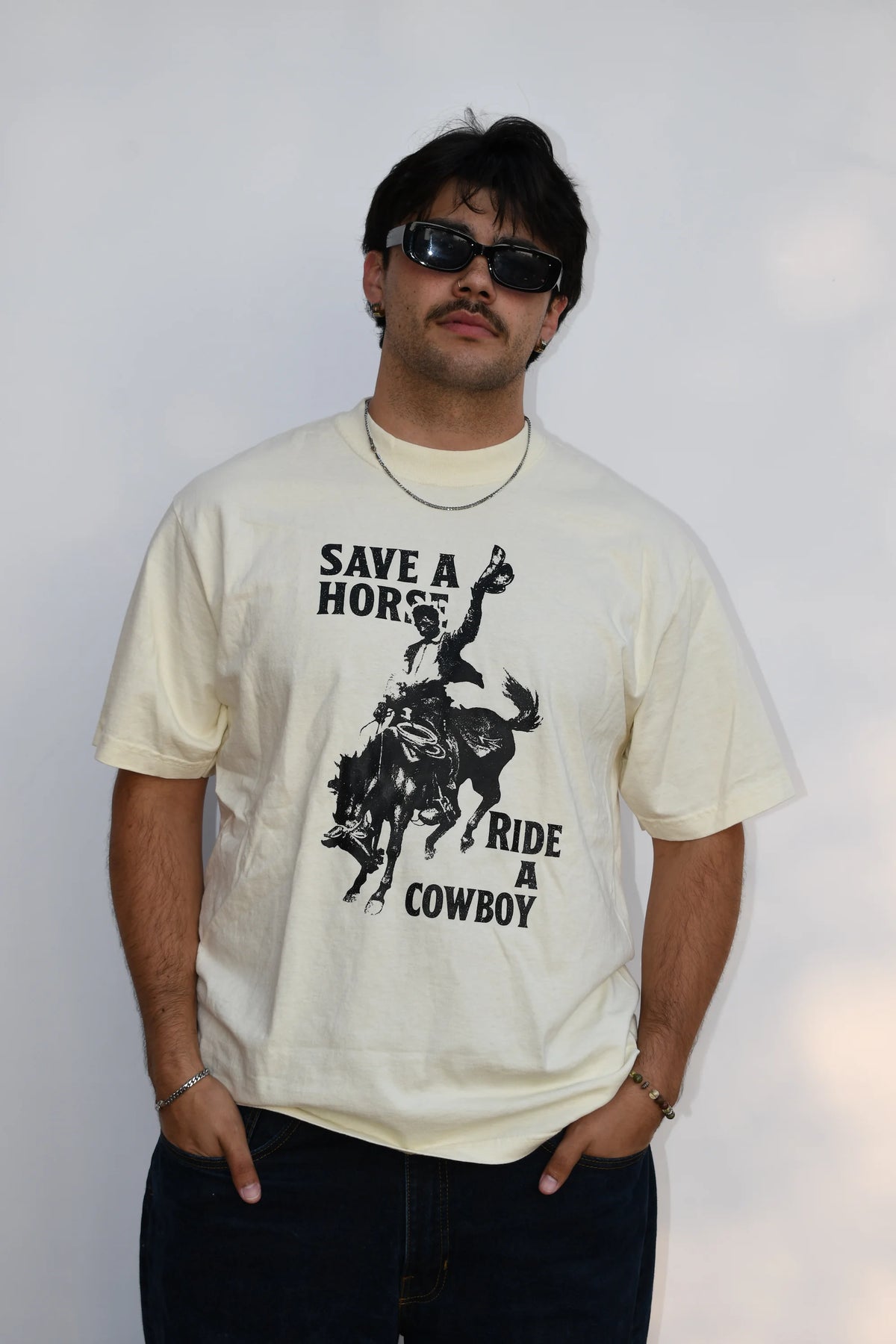 Model wearing a Common Saints cream graphic T-shirt with cowboy on a bucking horse and “Save a Horse Ride a Cowboy” text
