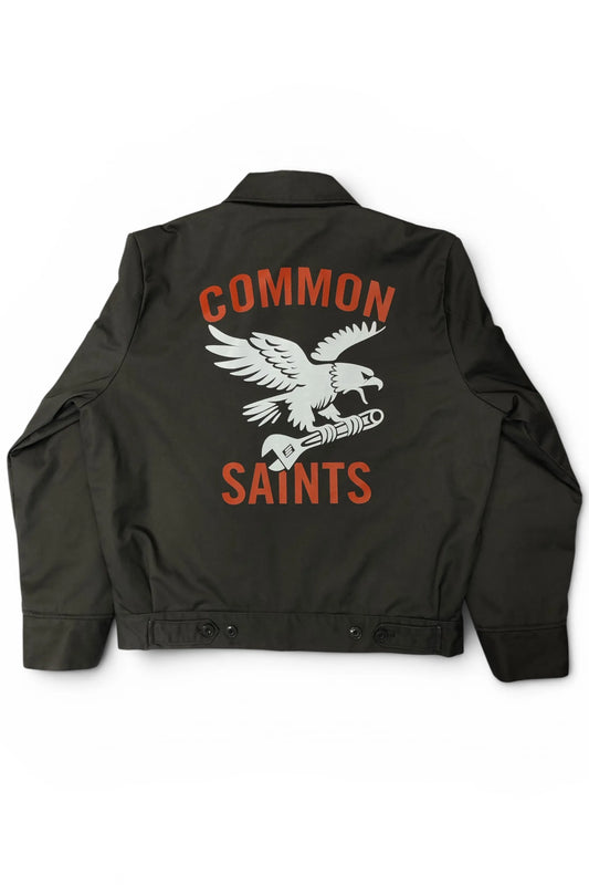 Common Saints dark gray mechanic jacket with large white eagle-and-wrench back graphic and red “Common Saints” lettering