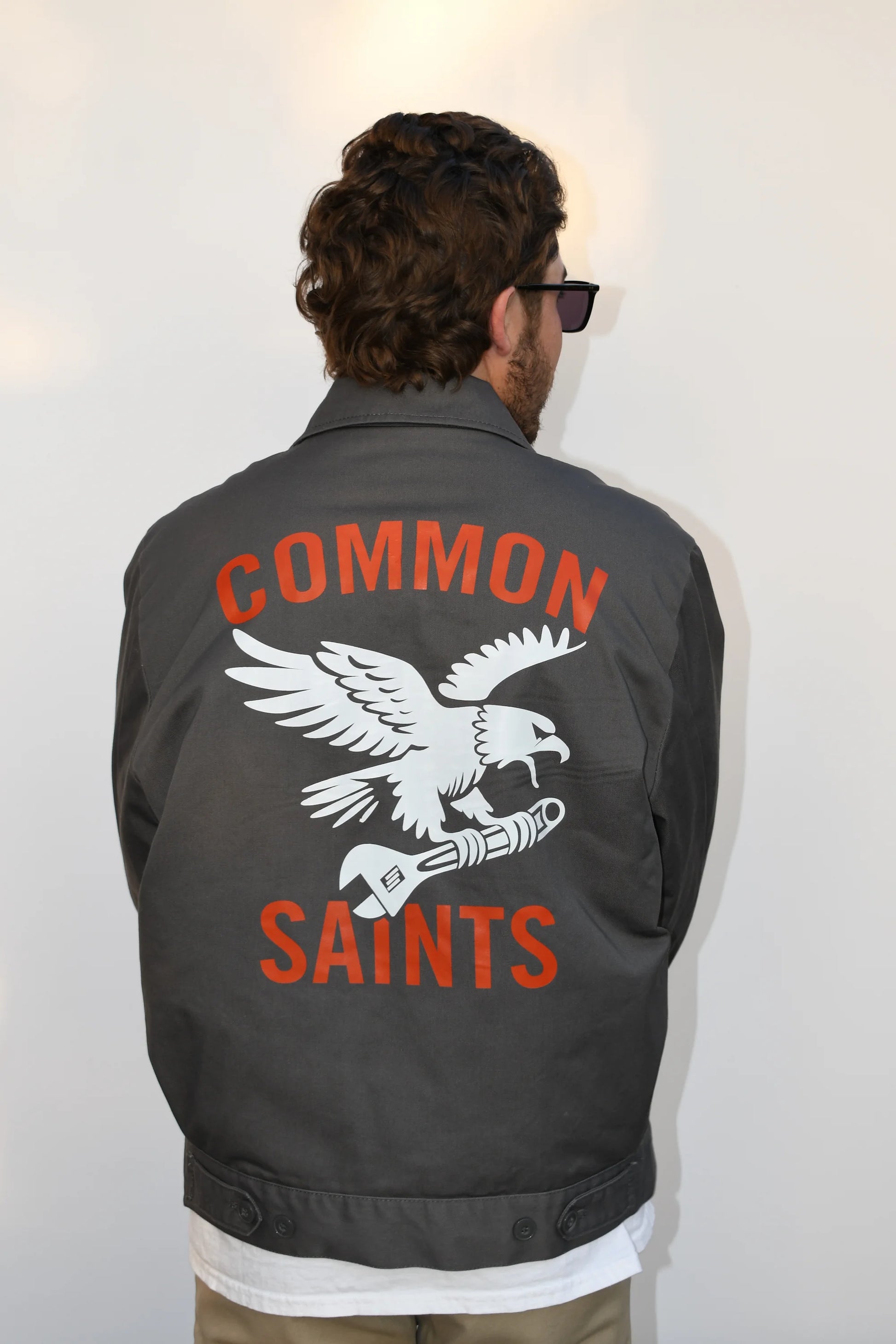 Back view of a model wearing the Common Saints dark gray mechanic jacket with white eagle-and-wrench graphic and Red logo text