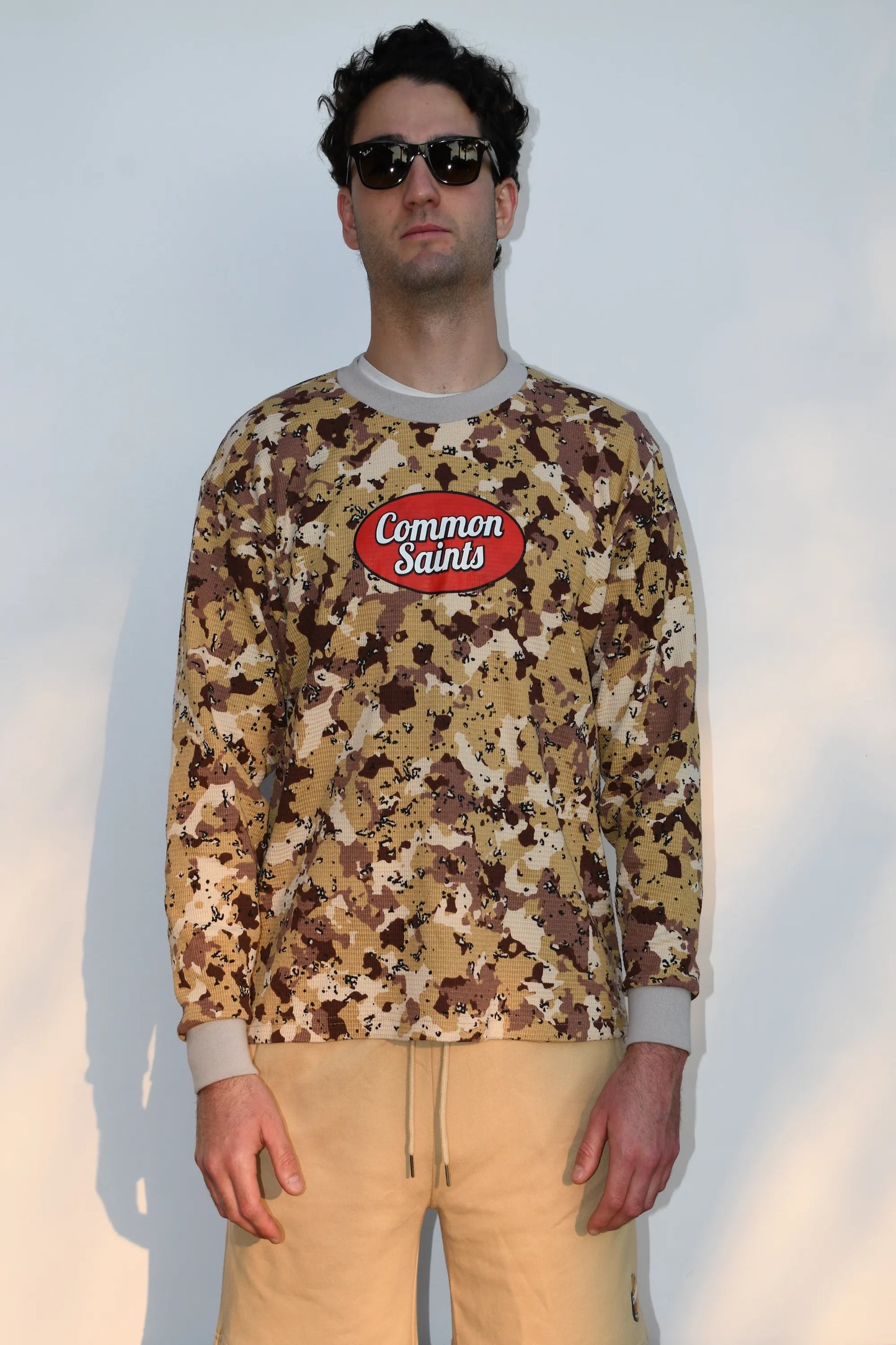 Model wearing a Common Saints desert camouflage long-sleeve crewneck with red oval chest logo and gray ribbed trim