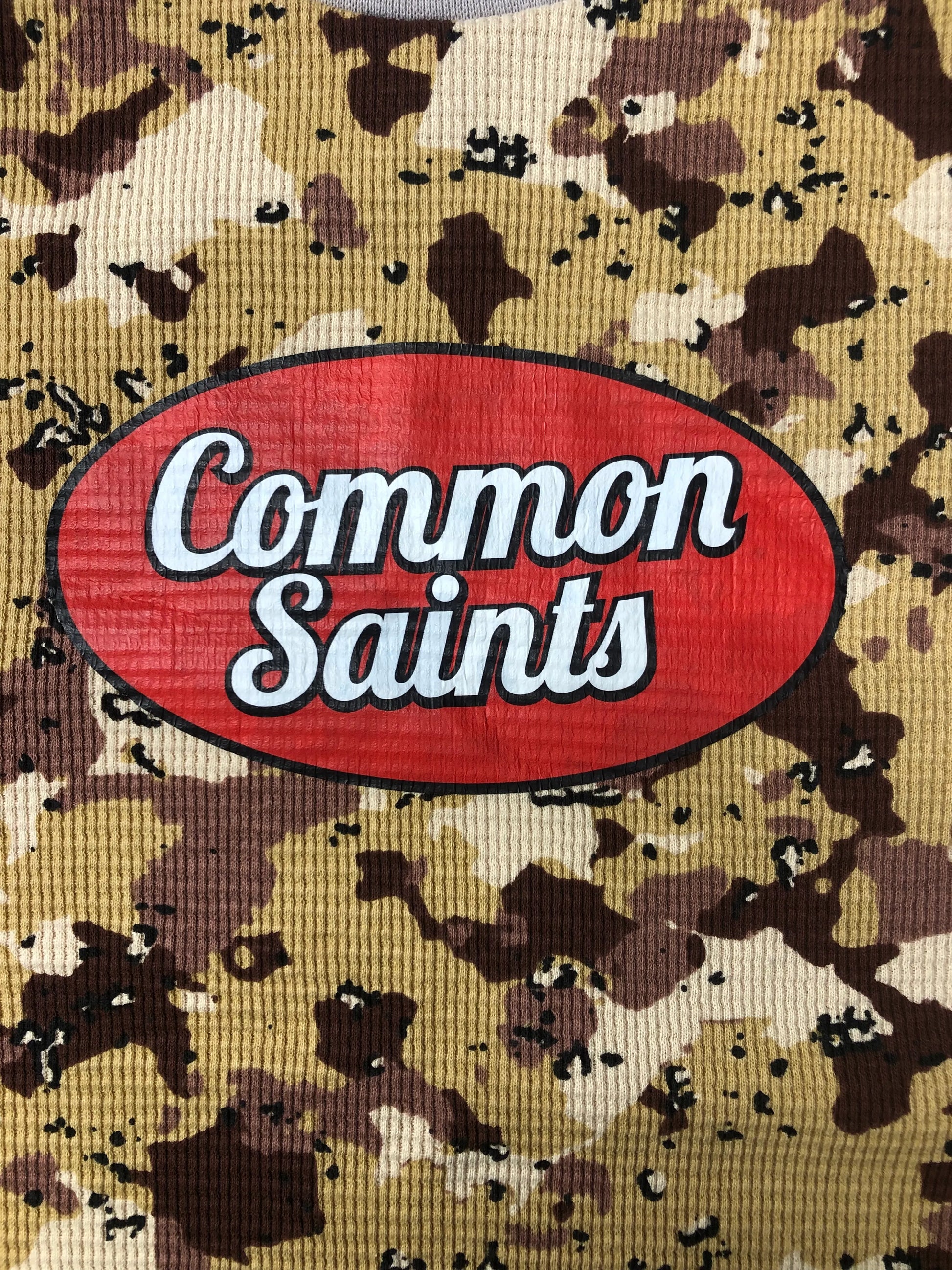 Close-up of the red oval Common Saints logo in white script on tan, brown, and beige desert camouflage fabric