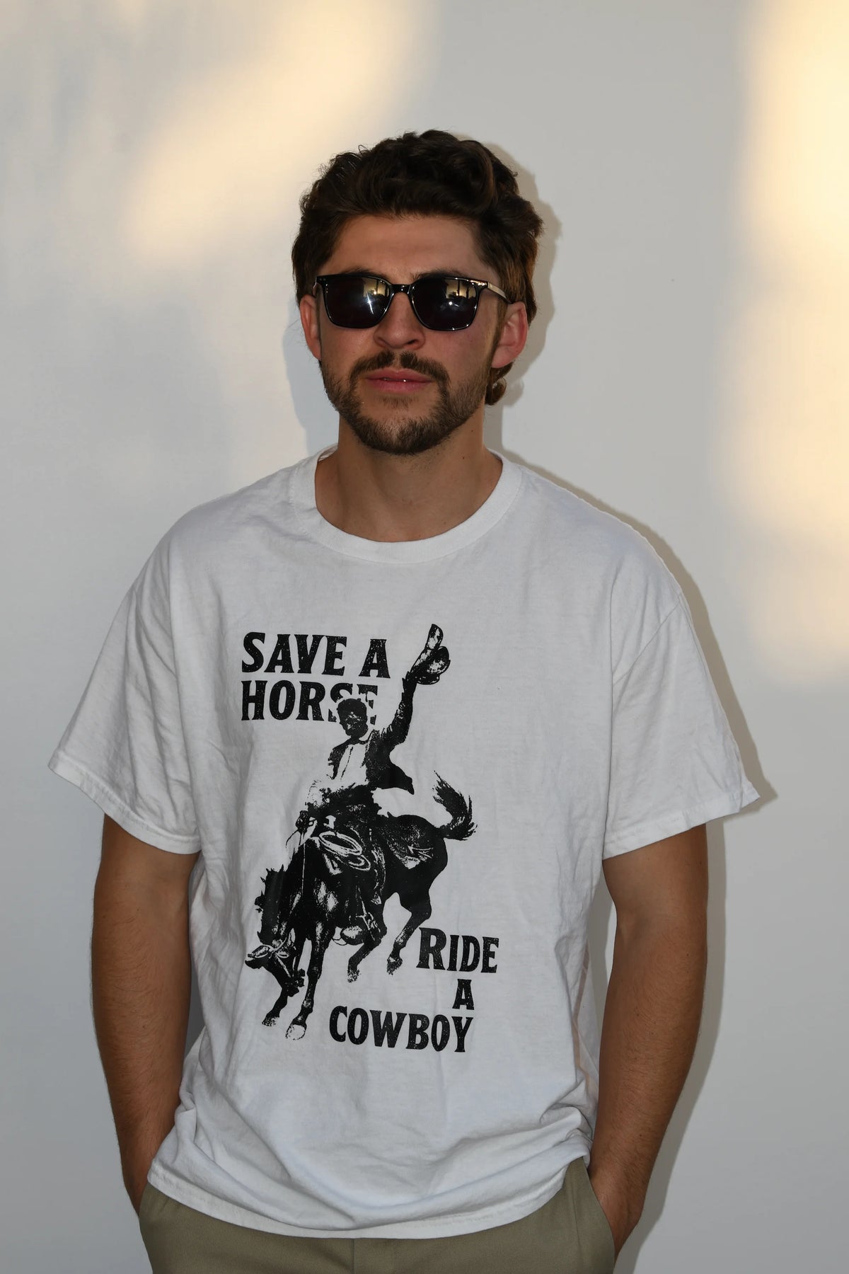 Angled front view of a model wearing a white Common Saints cowboy graphic T-shirt with “Save a Horse Ride a Cowboy” text