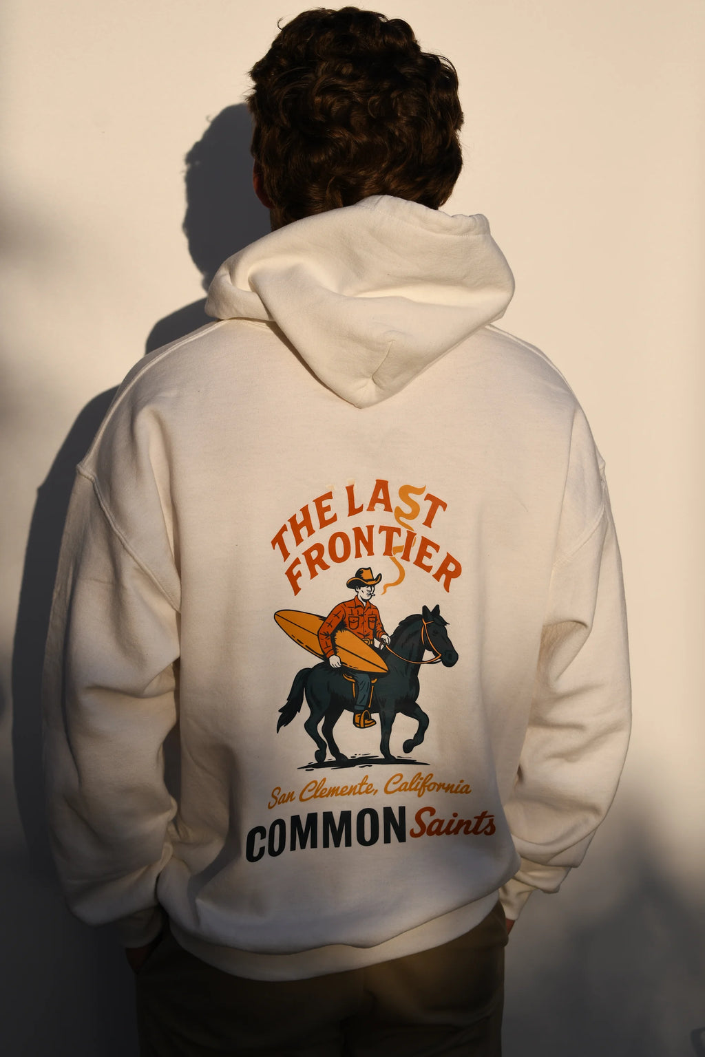 Model wearing a Common Saints white Last Frontier hoodie with cowboy, black horse, surfboard, and San Clemente back graphic