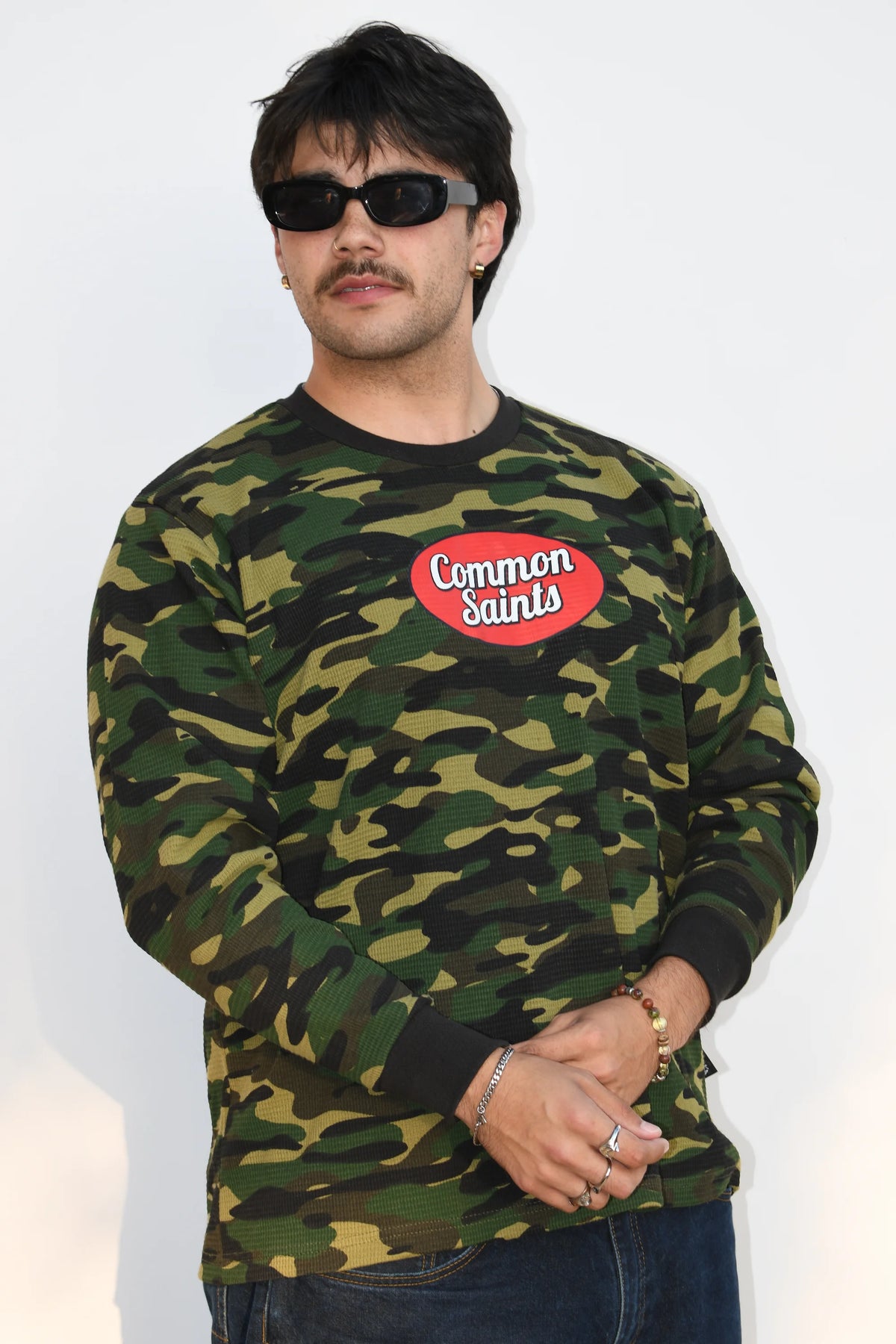 Model wearing the Common Saints Woodland Camo Heavyweight Thermal with red oval chest logo and black ribbed trim, angled front view
