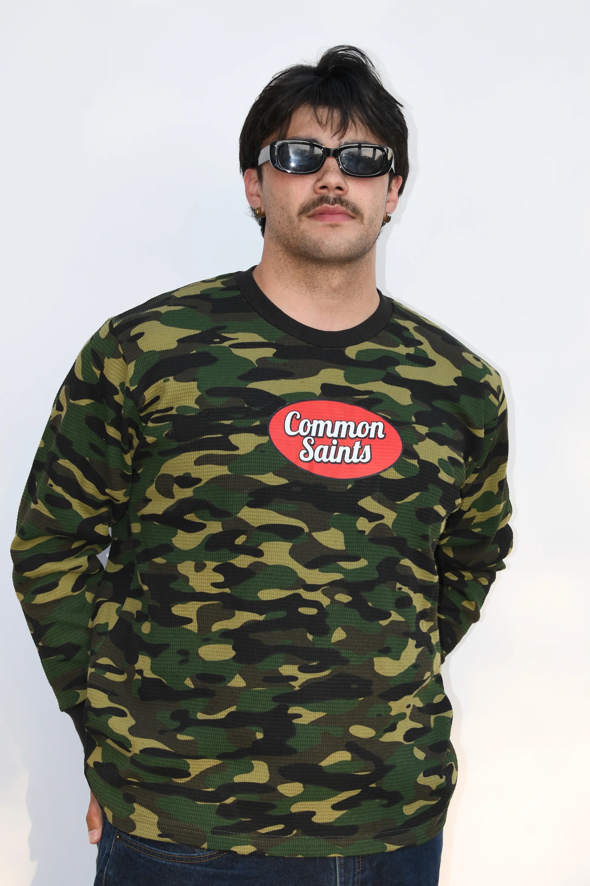 Front view of a model wearing the Common Saints Woodland Camo Heavyweight Thermal with red oval logo and black ribbed trim